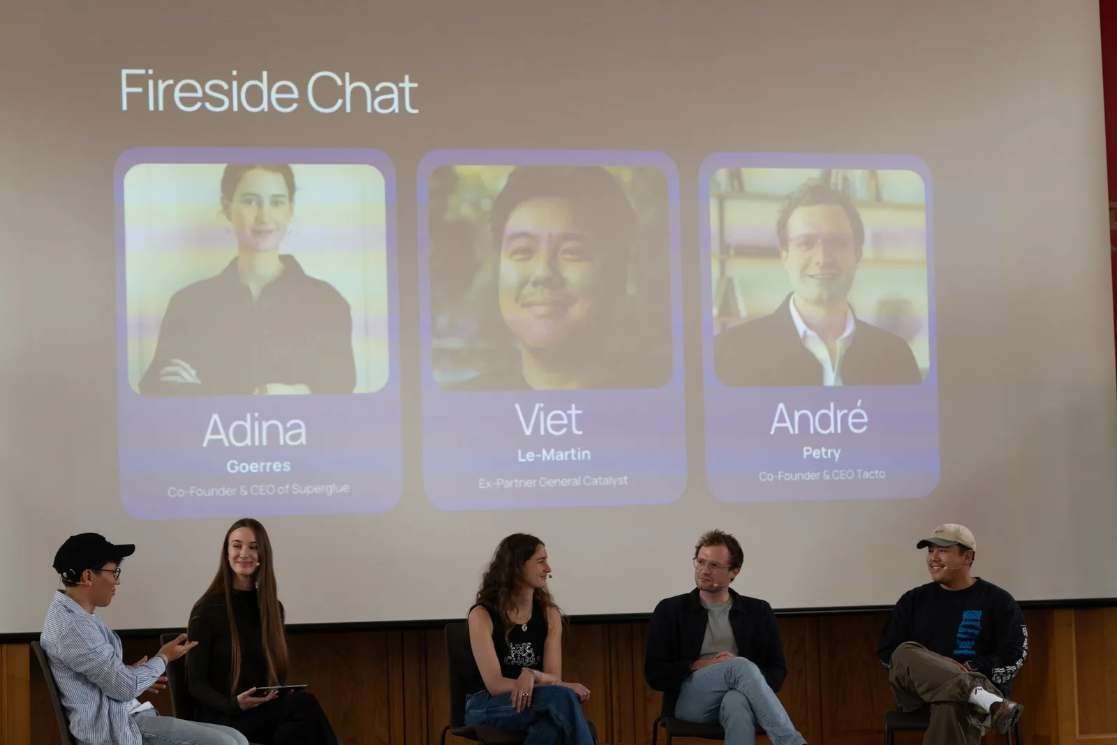 Five people seated on a stage during a fireside chat, with a slide behind them introducing the three guest speakers.