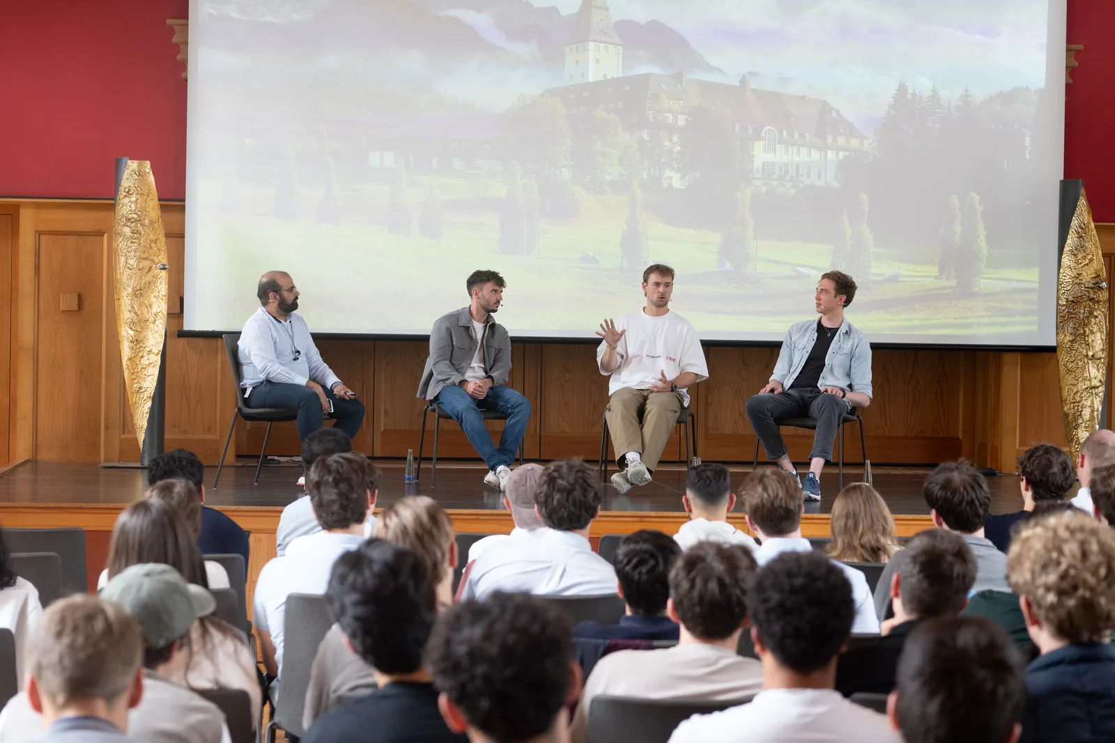 Four people seated on a stage in front of a large projected image of Schloss Elmau, speaking to a seated audience.
